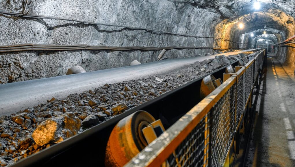 Aura-Minerals-Aktie-Starke-Dividenden-wachsendes-Geschaeft.jpeg Underground mine tunnel with a conveyor belt carrying rocks, rocky walls, and safety railing along the path.