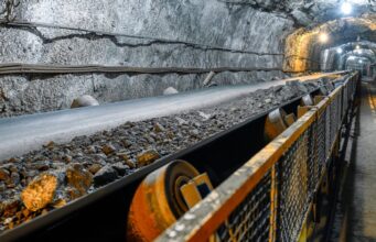 Aura Minerals-Aktie: Starke Dividenden, wachsendes Geschäft Underground mine tunnel with a conveyor belt carrying rocks, rocky walls, and safety railing along the path.