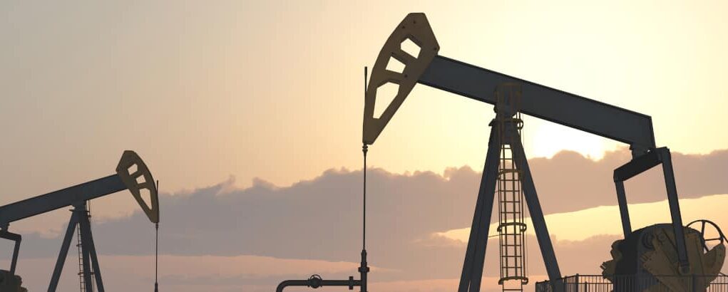 Two oil pumpjacks silhouetted against a warm sunset sky in an oil field at dusk.