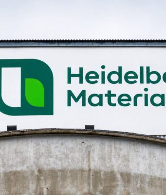 Heidelberg Materials-Aktie -14% im Juniverlauf Large Heidelberg Materials sign with green logo on a white panel against a blue industrial building exterior.
