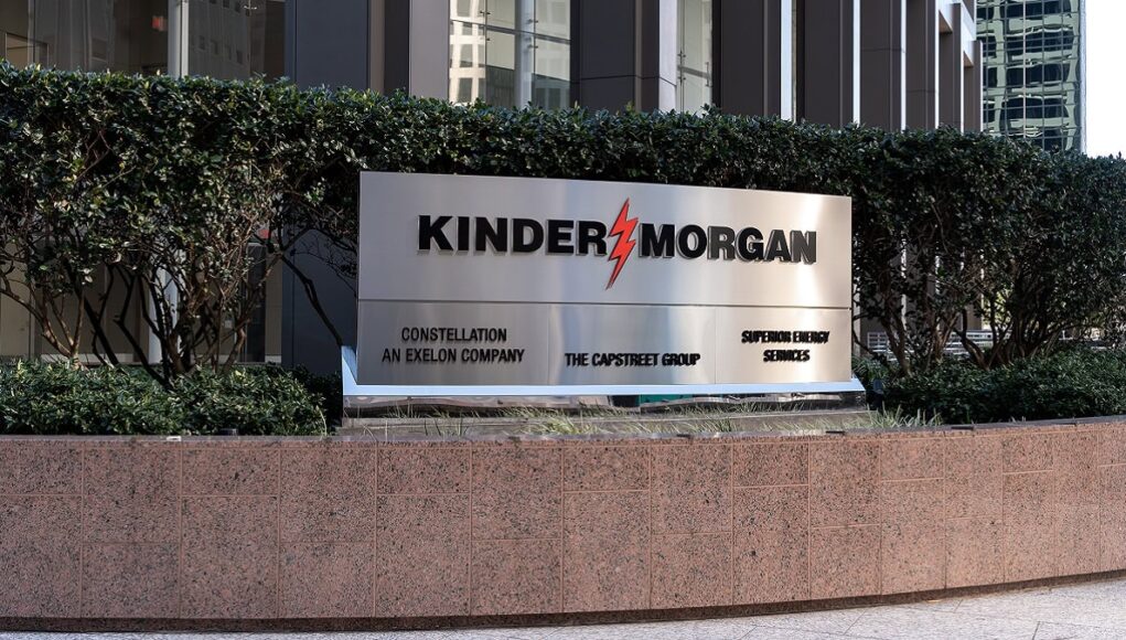 Exterior sign reading KINDER MORGAN with a red lightning bolt logo, set in front of hedges and a stone planter in a business district.