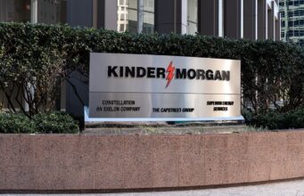 Kinder Morgan-Aktie: Gewinn und Dividende hoch Exterior sign reading KINDER MORGAN with a red lightning bolt logo, set in front of hedges and a stone planter in a business district.