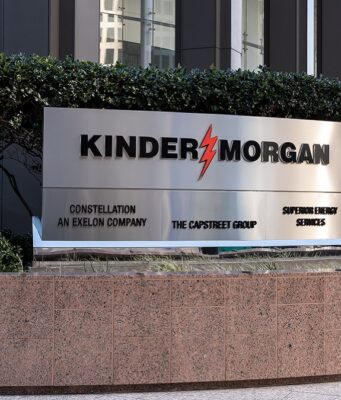 Kinder Morgan-Aktie: Gewinn und Dividende hoch Exterior sign reading KINDER MORGAN with a red lightning bolt logo, set in front of hedges and a stone planter in a business district.