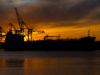 DAX jetzt: Rekordhoch in Reichweite dank sinkender Ölpreise Silhouette of a cargo ship with gantry cranes at a sunset harbor; orange sky and water reflections dominate the scene.