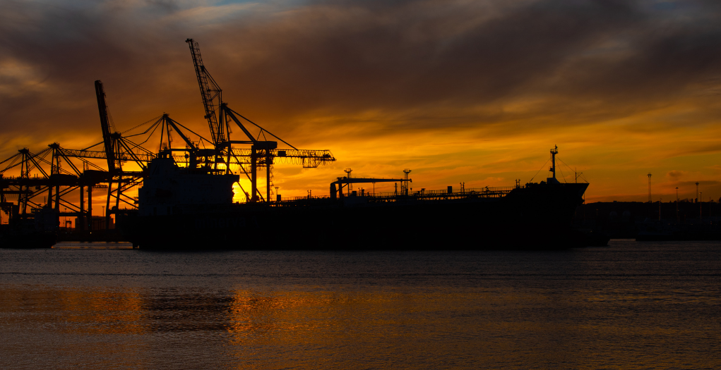 Silhouette of a cargo ship with gantry cranes at a sunset harbor; orange sky and water reflections dominate the scene.