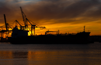 DAX jetzt: Rekordhoch in Reichweite dank sinkender Ölpreise Silhouette of a cargo ship with gantry cranes at a sunset harbor; orange sky and water reflections dominate the scene.