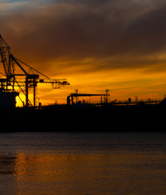DAX jetzt: Rekordhoch in Reichweite dank sinkender Ölpreise Silhouette of a cargo ship with gantry cranes at a sunset harbor; orange sky and water reflections dominate the scene.