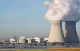 Cameco-Aktie: Diese Kursziele lassen Anleger aufhorchen Nuclear power plant with two large cooling towers releasing steam, set near a waterway with industrial buildings in the background.