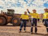 Newmont-Aktie: Startet nun das große Comeback? Three construction workers in yellow safety vests and hard hats stand on a gravel site, with a large yellow dump truck behind them and rocky hills in the distance.