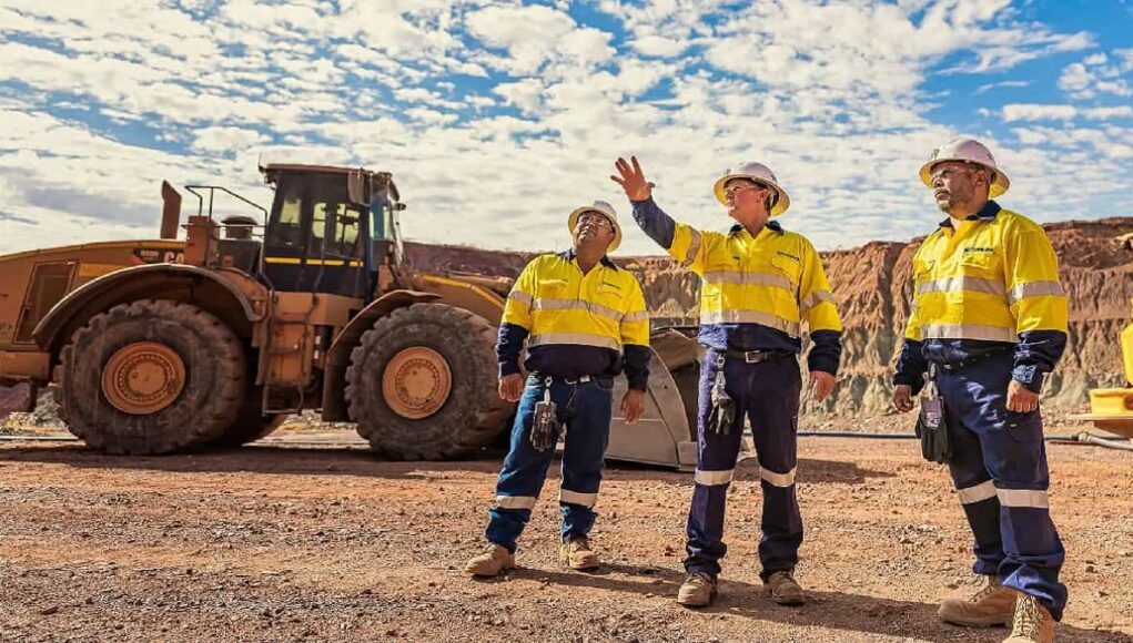 Three construction workers in yellow safety vests and hard hats stand on a gravel site, with a large yellow dump truck behind them and rocky hills in the distance.