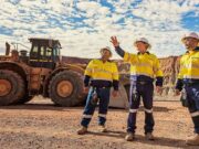 Newmont-Aktie: Startet nun das große Comeback? Three construction workers in yellow safety vests and hard hats stand on a gravel site, with a large yellow dump truck behind them and rocky hills in the distance.