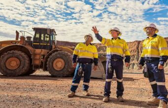 Newmont-Aktie: Startet nun das große Comeback? Three construction workers in yellow safety vests and hard hats stand on a gravel site, with a large yellow dump truck behind them and rocky hills in the distance.