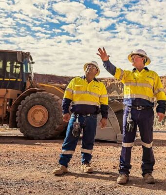 Newmont-Aktie: Startet nun das große Comeback? Three construction workers in yellow safety vests and hard hats stand on a gravel site, with a large yellow dump truck behind them and rocky hills in the distance.