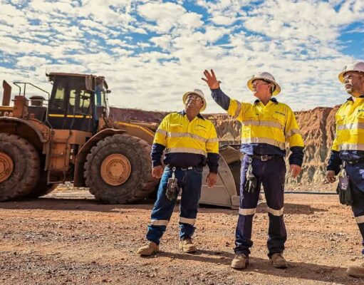 Newmont-Aktie: Startet nun das große Comeback? Three construction workers in yellow safety vests and hard hats stand on a gravel site, with a large yellow dump truck behind them and rocky hills in the distance.