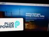 Plug Power-Aktie: Wo könnte ein erster Zielkurs liegen? Foreground: white smartphone showing the Plug Power logo in dark blue and teal, with a blurred blue-toned monitor displaying a hydrogen energy webpage in the background.