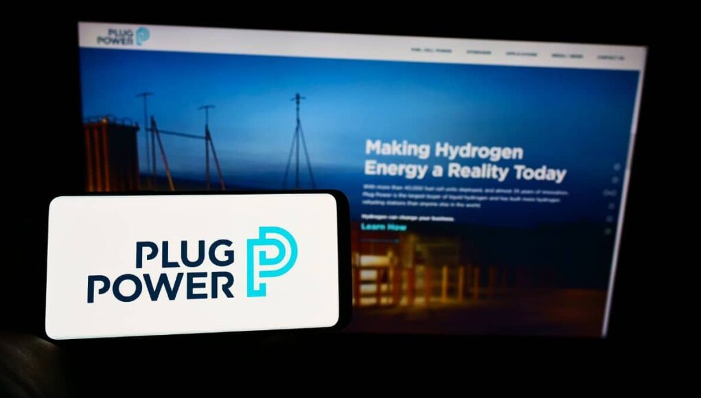 Foreground: white smartphone showing the Plug Power logo in dark blue and teal, with a blurred blue-toned monitor displaying a hydrogen energy webpage in the background.