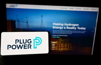 Plug Power-Aktie: Wo könnte ein erster Zielkurs liegen? Foreground: white smartphone showing the Plug Power logo in dark blue and teal, with a blurred blue-toned monitor displaying a hydrogen energy webpage in the background.