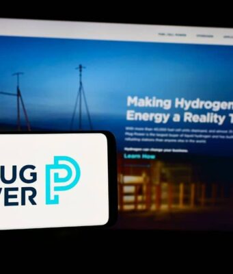 Plug Power-Aktie: Wo könnte ein erster Zielkurs liegen? Foreground: white smartphone showing the Plug Power logo in dark blue and teal, with a blurred blue-toned monitor displaying a hydrogen energy webpage in the background.