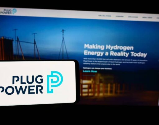 Plug Power-Aktie: Wo könnte ein erster Zielkurs liegen? Foreground: white smartphone showing the Plug Power logo in dark blue and teal, with a blurred blue-toned monitor displaying a hydrogen energy webpage in the background.
