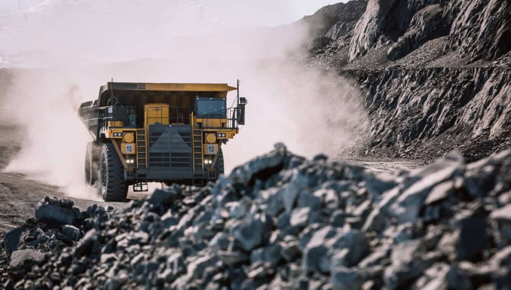 Rio-Tinto-Aktie-der-Kupferpreis-in-luftigen-Hoehen.jpeg Yellow mining dump truck drives through a dusty rocky quarry with steep rock faces on either side