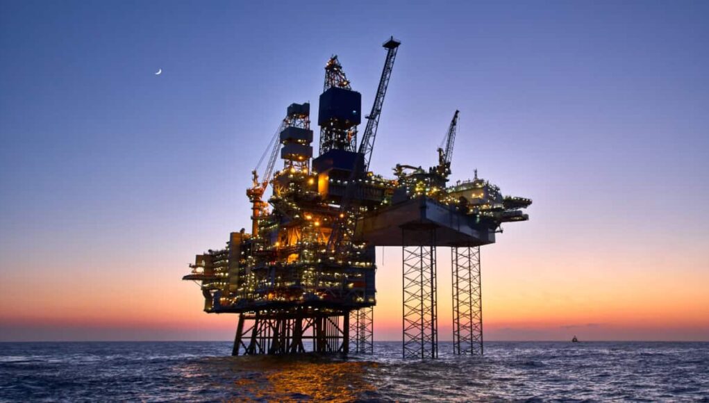 Chevron-Aktie-Oel-Boom-trifft-auf-KI-Fantasie.jpeg Offshore oil platform lit up at dusk, standing on stilts above calm ocean with a crescent moon in the sky.
