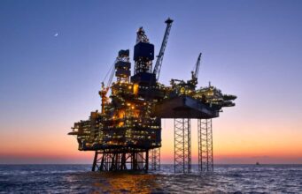Chevron-Aktie: Öl-Boom trifft auf KI-Fantasie Offshore oil platform lit up at dusk, standing on stilts above calm ocean with a crescent moon in the sky.