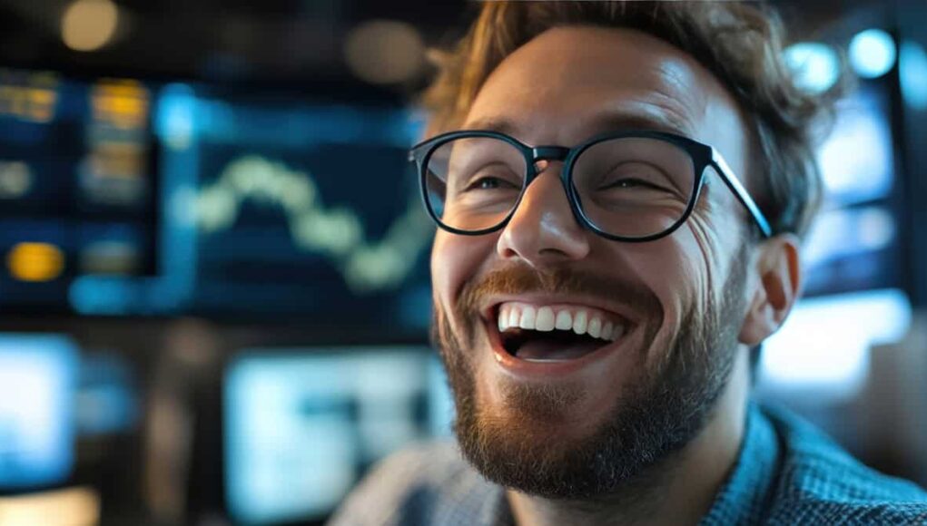 Close-up of a smiling man with a beard and black-rimmed glasses in a high-tech workspace.