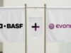 Evonik-Aktie: Kommt jetzt doch eine schnelle Übernahme? Three white banners on a metal stand showing BASE on the left, a central banner with a black plus and purple vertical bar, and the EVONIK logo on the right.