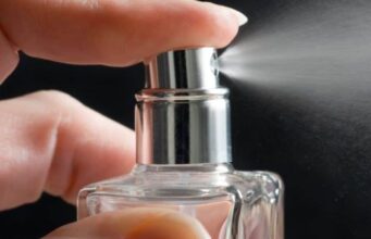 Symrise-Aktie +40% seit Jahresanfang: Was könnte jetzt ratsam sein? Close-up of a hand pressing a perfume spray nozzle and releasing mist from a glass bottle